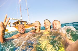 young multiracial friends taking selfie and swimming on sailing boat sea trip - rich happy guys and girls having fun in summer party day - exclusive vacation concept - bright afternoon warm filter