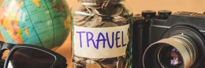 coins in glass jar with accessories of traveler on wooden table. saving money for travel concept