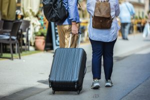 Elderly married couple walking through city center with suitcases. They are having hats and backpacks on.