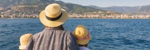 a mother and two children wearing straw hats seen from behind gazing at a scenic coastal city from a boat