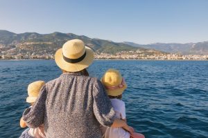 a mother and two children wearing straw hats seen from behind gazing at a scenic coastal city from a boat