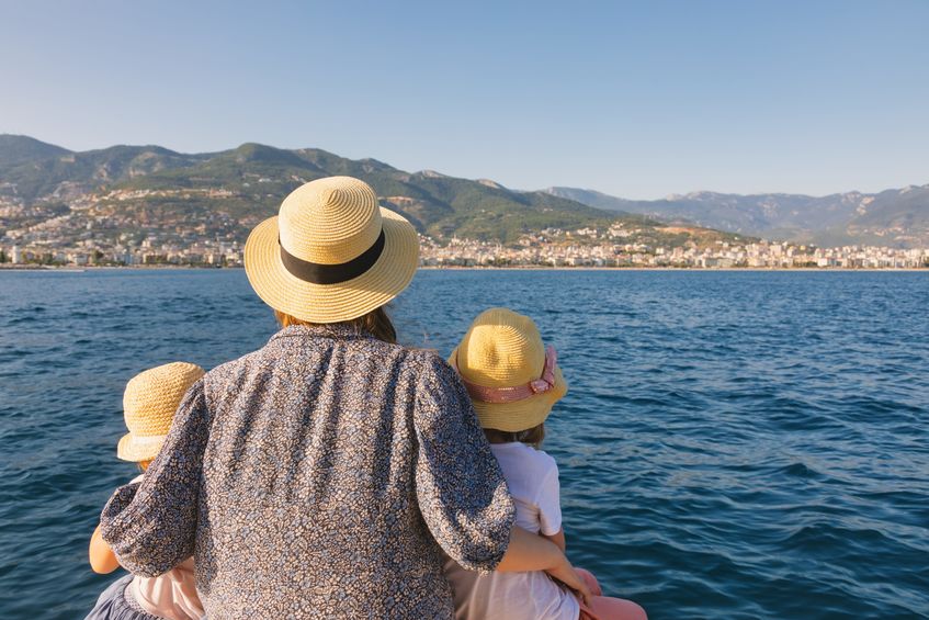 a mother and two children wearing straw hats seen from behind gazing at a scenic coastal city from a boat